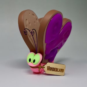 Puede incluir: Una caprichosa figura de mariposa rosa con ojos verdes y alas moradas. Las alas tienen forma de rebanadas de pan con corteza marrón e interior beige. Una etiqueta de madera dice "Wonderland".