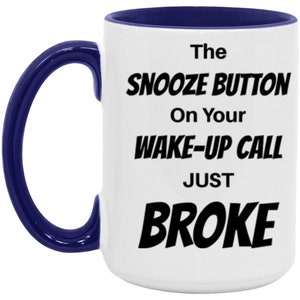 May include: White ceramic coffee mug with a blue rim and handle. The mug has black text that reads "The Snooze Button On Your Wake-Up Call Just Broke".