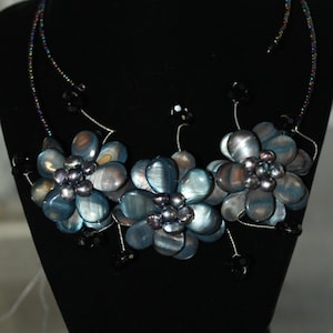 May include: A silver necklace with three blue iridescent flower-shaped pendants and black beads. The necklace is on a black velvet display stand.