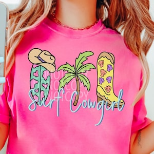 May include: A hot pink t-shirt with a graphic that says "Surf Cowgirl". The graphic includes a surfboard with hearts, a cowboy hat, a palm tree, and a cowboy boot with seashells.
