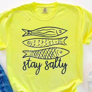 May include: A bright yellow t-shirt with three stylized fish in black and the words "stay salty" in a script font. The shirt is paired with denim shorts and white sandals.