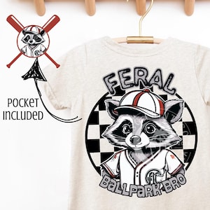 Op de afbeelding: Een beige T-shirt met een afbeelding van een wasbeer die een baseballpet en een shirt draagt. De wasbeer bevindt zich in een cirkel met een geruit patroon. De tekst "FERAL BALLPARK BRO" staat onder de wasbeer. Het shirt heeft een zakje aan de voorkant.