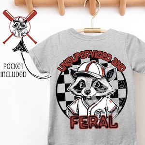 Op de afbeelding: Grijs T-shirt met korte mouwen met een afbeelding van een wasbeer die een baseballpet en een shirt draagt. De wasbeer bevindt zich in een cirkel met een geruite achtergrond. De tekst "Unsupervised and Feral" is op het T-shirt gedrukt.