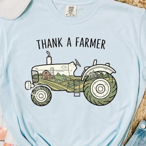 Op de afbeelding: Een lichtblauw t-shirt met de tekst "THANK A FARMER" boven een tractorillustratie. Het tractorontwerp toont een boerderij met een schuur, velden en een boerderij. Het shirt wordt getoond met een spijkerbroek en een witte pet.