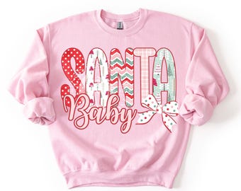 May include: A light pink sweatshirt with the words "SANTA BABY" in a festive design. The letters are filled with patterns like polka dots, zigzags, and Christmas trees. A bow with polka dots is at the bottom right.