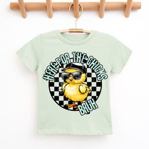 Puede incluir: Camiseta verde claro con un gr&aacute;fico de un pollito amarillo con gafas de sol y una gorra negra. El texto "HERE FOR THE CHICKS BRUH" est&aacute; impreso alrededor del pollito sobre un fondo a cuadros.