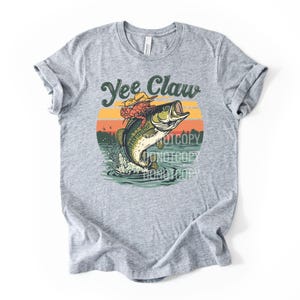 May include: A gray t-shirt featuring a graphic of a fish jumping out of water with a crab wearing a cowboy hat. The text "Yee Claw" is above the graphic, with a sunset background.