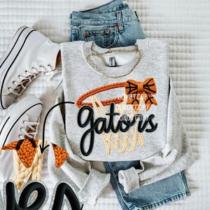 May include: A light gray sweatshirt with a basketball hoop design and the word "Gators" in black script. The design includes orange and beige accents. The sweatshirt is paired with blue jeans and white sneakers.