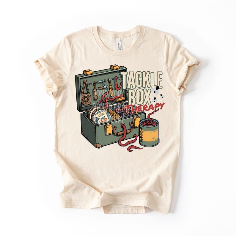Op de afbeelding: Cr&egrave;mekleurig T-shirt met een afbeelding van een groene tacklebox met visgerei en de tekst "Tackle Box Therapy". Een blikje aas en rode wormen zijn ook afgebeeld. Het shirt is gemaakt van een zacht materiaal.