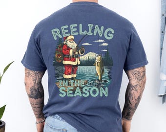 May include: Navy blue t-shirt with a graphic of Santa Claus fishing, with the text "Reeling in the Season." Santa is wearing a red suit and holding a fishing rod. A fish is jumping out of the water. The shirt is made of Comfort Colors denim.