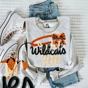 May include: A light gray sweatshirt with a basketball hoop design and the word "Wildcats" in black. The sweatshirt is paired with blue jeans and white sneakers, suggesting a sporty theme.