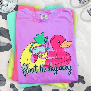May include: A stack of colorful t-shirts in pink, green, and yellow. The top shirt is lavender and features a graphic of a yellow pineapple wearing goggles and a pink inflatable duck with the text "float the day away".