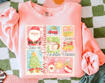 May include: A light pink sweatshirt featuring a Christmas-themed graphic. The graphic includes postage stamp-style images of Santa Claus, a car, mittens, a mug of hot cocoa, a Christmas tree, and ornaments. The words "Merry Christmas", "Snow", "Joy", and "Santa Claus" are also included.