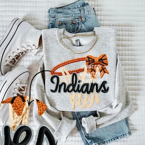 May include: A light gray sweatshirt with a basketball hoop design in orange and tan, and the word "Indians" in black script. The sweatshirt is paired with blue jeans and white high-top sneakers.