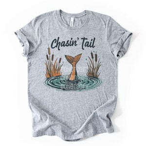 May include: A heather gray t-shirt featuring the phrase "Chasin' Tail" above a graphic of a mermaid tail emerging from water. The design includes cattails and water ripples, creating a nature-inspired theme.