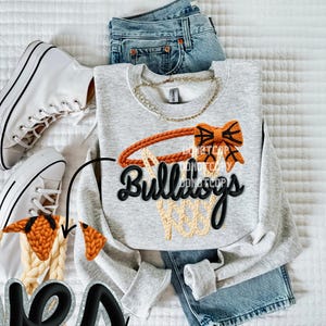 May include: A light gray sweatshirt with a basketball hoop design. The design includes an orange hoop and bow, with the word "Bulldogs" in black script. The sweatshirt is paired with blue jeans and white sneakers.