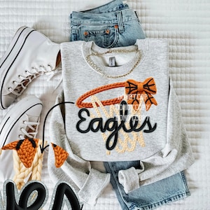 May include: A light gray sweatshirt with a basketball hoop design. The hoop is orange and tan, with the word "Eagles" in black script. White high-top sneakers and blue jeans are also in the image.