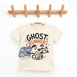 以下が含まれることがあります： クリーム色の半袖Tシャツ。幽霊とベースにスライディングする野球選手のグラフィックが描かれています。黒とオレンジの文字で「GHOST RUNNERS CLUB」と書かれています。CSG DESIGNSという小さなロゴも確認できます。