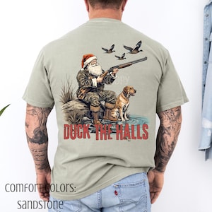 Op de afbeelding: Zandkleurig t-shirt met een afbeelding van de Kerstman met een geweer, vergezeld van een hond en eenden. De tekst "DUCK THE HALLS" is in het rood gedrukt. Het shirt maakt deel uit van de Comfort Colors collectie.