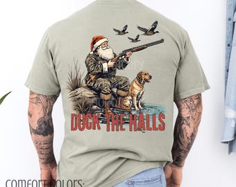 May include: Sandstone-colored t-shirt featuring a graphic of Santa Claus aiming a shotgun, accompanied by a dog and ducks. The text "DUCK THE HALLS" is printed in red. The shirt is part of the Comfort Colors collection.