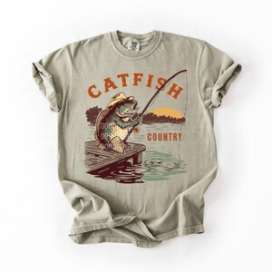 May include: A light green t-shirt with a graphic of a catfish wearing a cowboy hat fishing on a dock. The text "CATFISH COUNTRY" is above and below the image in orange. The shirt is made of a soft material.