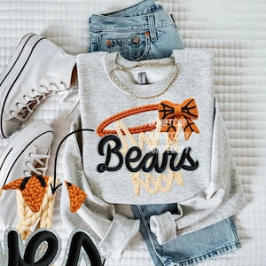 May include: A light gray sweatshirt with a basketball hoop design and the word "Bears" in black. The hoop is orange and the net is beige. White sneakers and blue jeans are also in the image.