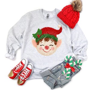 Puede incluir: Una sudadera gris claro con un diseño bordado de cara de elfo, un gorro rojo tejido con un pompón de piel sintética, zapatillas rojas y pantalones cortos de mezclilla. La cara del elfo tiene un sombrero rojo, acebo verde y una expresión sonriente. El texto "SG DESIGN" está en el sombrero del elfo.