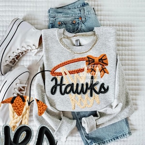 May include: A light gray sweatshirt with a basketball hoop design and the word "Hawks" in black. The hoop is orange and the net is beige. White sneakers and blue jeans are also in the image.