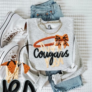 May include: A light gray sweatshirt with a basketball hoop design and the word "Cougars" in black. The hoop is orange and the net is beige. The sweatshirt is paired with blue jeans and white sneakers.