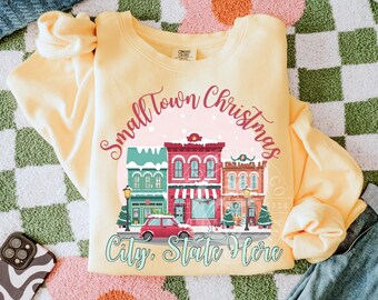 May include: A light yellow sweatshirt with a graphic that reads "Small Town Christmas" above a row of colorful buildings and a red car. The text "City, State Here" is below the buildings.