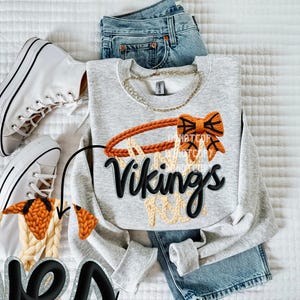 May include: A light gray sweatshirt with a basketball hoop design. The hoop is orange and tan, with the word "Vikings" in black script. The sweatshirt is paired with blue jeans and white sneakers.