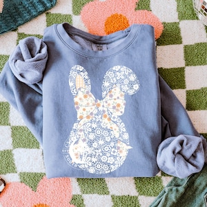 Puede incluir: Una sudadera azul con un diseño de conejo floral blanco y el texto "Est. South Wilds" en un círculo.
