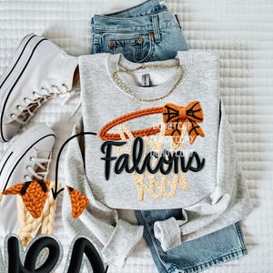 May include: A light gray sweatshirt with a basketball hoop design and the word "Falcons" in black. The design includes an orange hoop and a bow. The sweatshirt is paired with blue jeans and white sneakers.