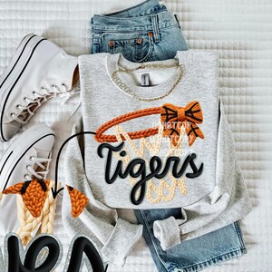 May include: A light gray sweatshirt with a basketball hoop design in orange and tan, and the word "Tigers" in black. The sweatshirt is paired with blue jeans and white sneakers.