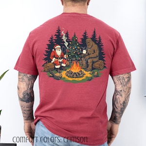 Könnte beinhalten: Karmesinrotes T-Shirt mit einer Grafik von Santa Claus und Bigfoot an einem Lagerfeuer mit einem geschmückten Weihnachtsbaum. Der Text "COMFORT COLORS: CRIMSON" befindet sich am unteren Rand des T-Shirts.