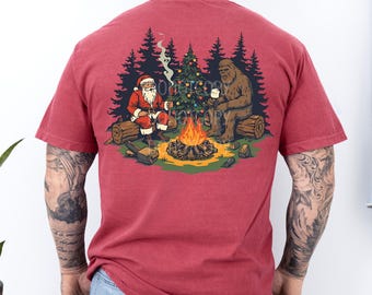 May include: A crimson red short-sleeve t-shirt featuring a graphic on the back of Santa Claus and Bigfoot sitting on logs by a campfire. They are roasting marshmallows in front of a decorated Christmas tree and dark pine forest background.