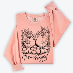 May include: A peach-colored sweatshirt with a black and white illustration of two chickens sitting on a nest of eggs. The text "Homestead Era" is printed below the chickens.