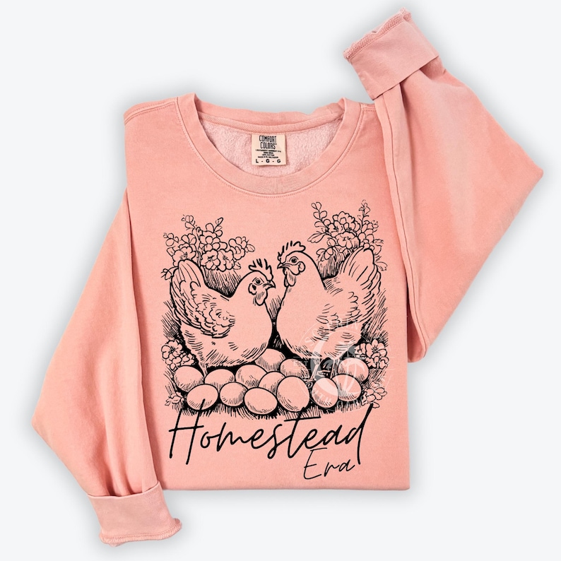 May include: A peach-colored sweatshirt with a black and white illustration of two chickens sitting on a nest of eggs. The text "Homestead Era" is printed below the chickens.
