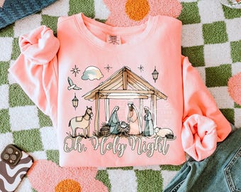 May include: A light pink sweatshirt with a graphic of the Nativity scene, including a stable, figures, and animals. The text "Oh. Holy Night" is below the graphic. The sweatshirt is on a green and white checkered surface.