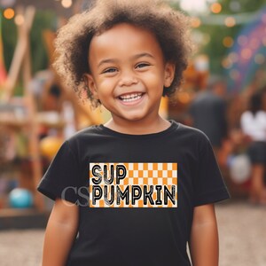 May include: A child wearing a black t-shirt with a checkered orange and white design and the text "Sup Pumpkin" in black.