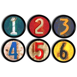 May include: Six round decorative magnets featuring the numbers one through six. Each number is in a different color, with a distressed, vintage look. The magnets have black borders and are arranged in two rows of three.