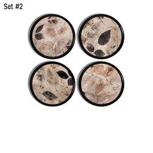May include: Set #2 consists of four circular coasters with black rims. Each coaster features a beige background decorated with a botanical print of dark brown and tan leaves, twigs, and foliage in a variety of shapes and sizes across the surface.