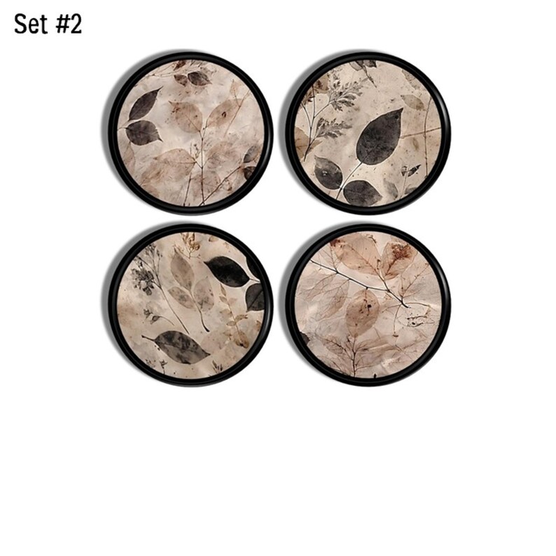 May include: Set #2 consists of four circular coasters with black rims. Each coaster features a beige background decorated with a botanical print of dark brown and tan leaves, twigs, and foliage in a variety of shapes and sizes across the surface.