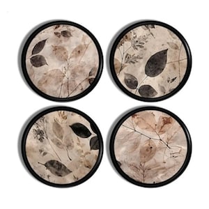 May include: A set of four circular coasters, each featuring a black frame and a beige background decorated with a botanical print of various dark brown and tan leaves and twigs. The designs on each coaster vary, showcasing different leaf arrangements.