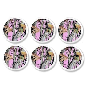 May include: Six round white-rimmed buttons with a pink camouflage pattern. The pattern includes tree branches, leaves, and a pink background. The buttons are arranged in two rows of three.