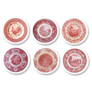 May include: Six round coasters with a white border, each featuring a different red and white toile design. The designs include a rooster, landscapes, and floral patterns. These coasters are perfect for protecting surfaces.