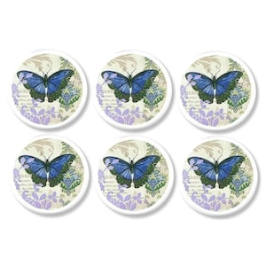 May include: Six round coasters with a butterfly design. Each coaster features a blue butterfly with green accents on a cream background with purple floral and text details. The coasters are arranged in two rows of three.