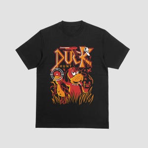May include: Black t-shirt with a cartoon illustration of a dog and duck in a hunting scene. The design features the text "DUCK HUNT" in orange and red, with a target reticle and a duck in the crosshairs.