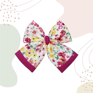 May include: A large fabric hair bow featuring a white background with a colourful floral pattern of pink, yellow, and orange flowers. The bow is cinched in the centre with a solid dark pink fabric knot and has solid dark pink borders on the ends.