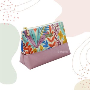 Puede incluir: Un estuche con cremallera que presenta un estampado floral y botánico con hojas vibrantes en naranja, azul y amarillo sobre un fondo crema. La sección inferior es de tela malva sólida. Incluye una pequeña etiqueta metálica con anjurisa.